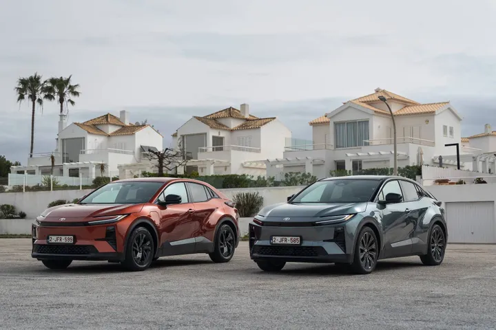 Foto di due esemplari di Toyota C-HR+, una rossa metallizzata e l'altra grigia metallizzata, fotografate frontalmente con una città marittima di sfondo