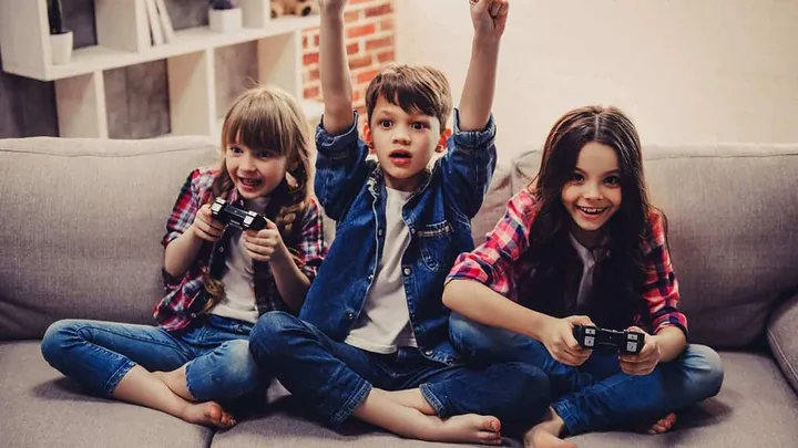 cheerful kids are sitting together on sofa at home girls are playing game console while boy is cheering for them