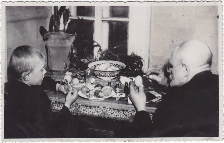 Fotonuotrauka. Grupinė. Senelis (dešinėje) su anūku (kairėje) dažo velykinius margučius prie uždaroje patalpoje esančio stalo. XX a. 3–4 deš. (Trakų muziejus, CC BY)